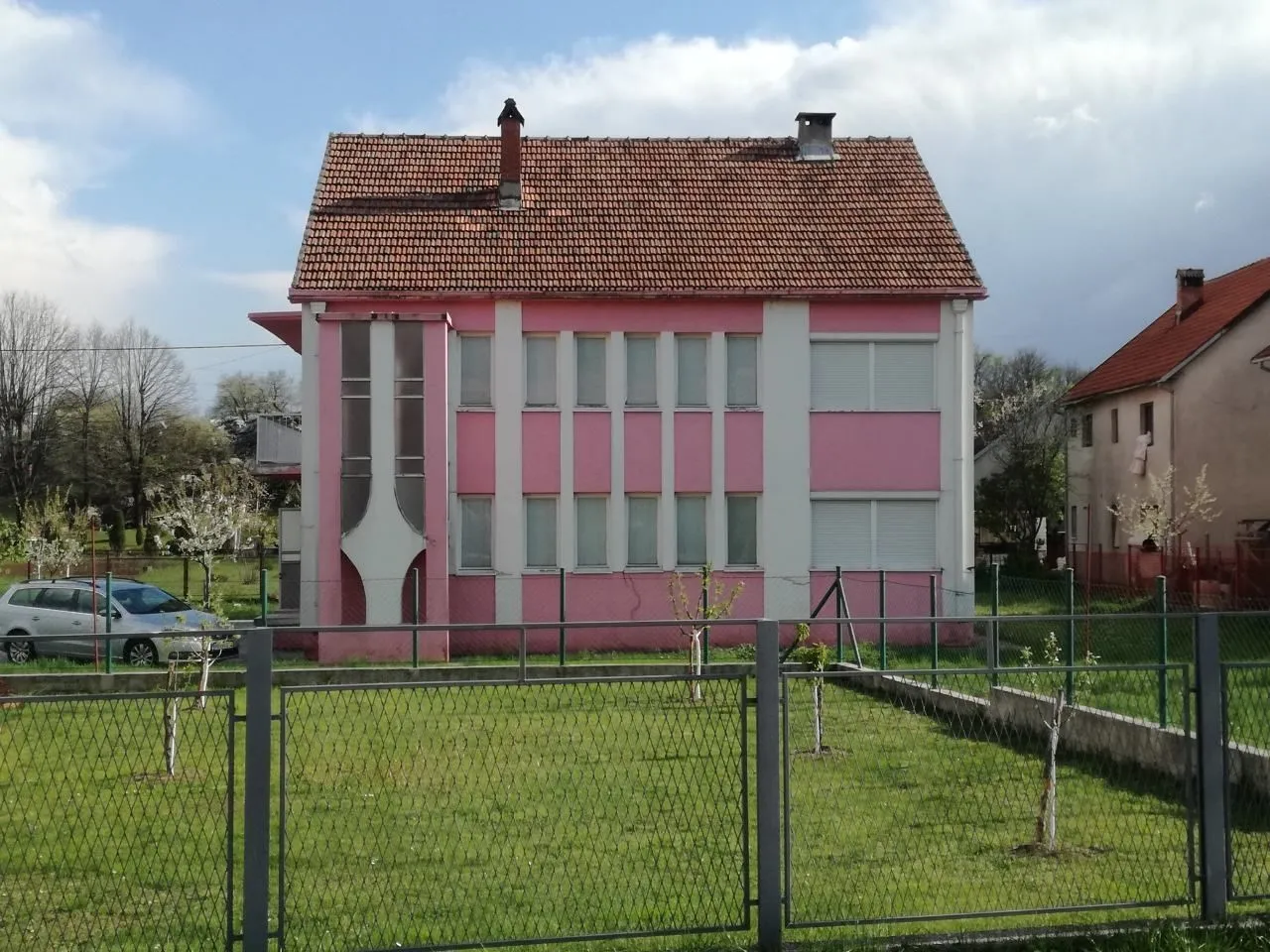 Dvospratna kuća sa roze fasadom i ograđenim dvorištem.