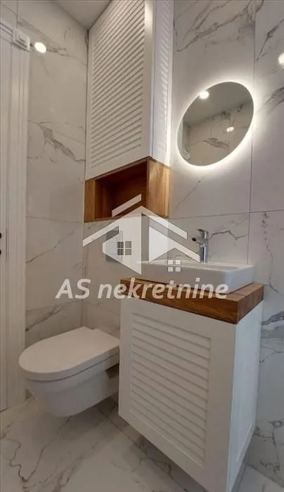 Moderno kupatilo sa belim pločicama, umivaonikom i ugrađenom toalet šoljom.