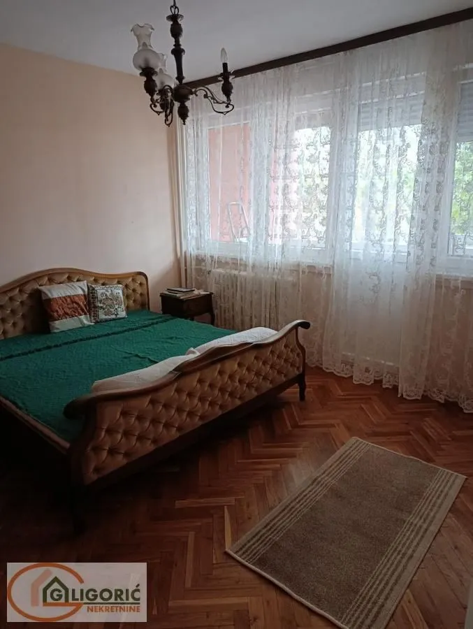 Spavaća soba sa bračnim krevetom, drvenim parketom i prozorima sa belim zavesama.