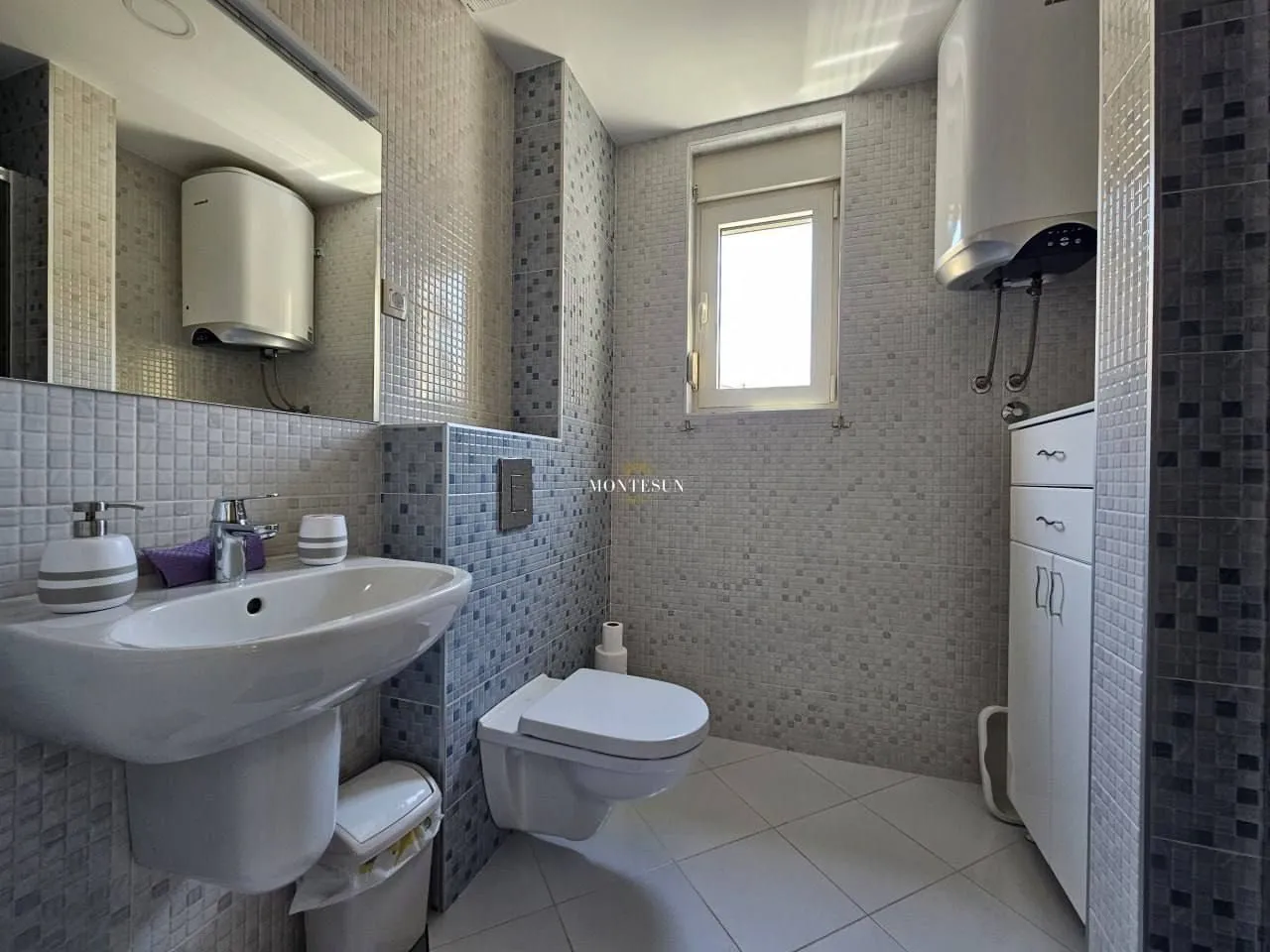 Small mosaic-tiled bathroom with a washbasin, toilet, and water heater.