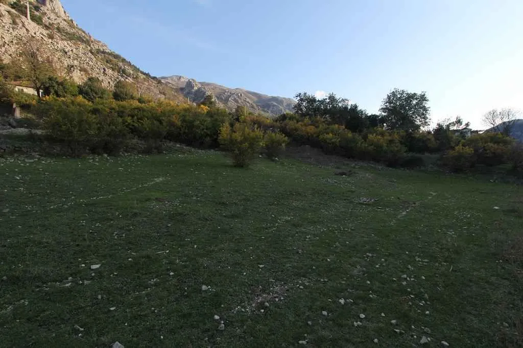 Prostran travnati plac okružen drvećem podno planine.