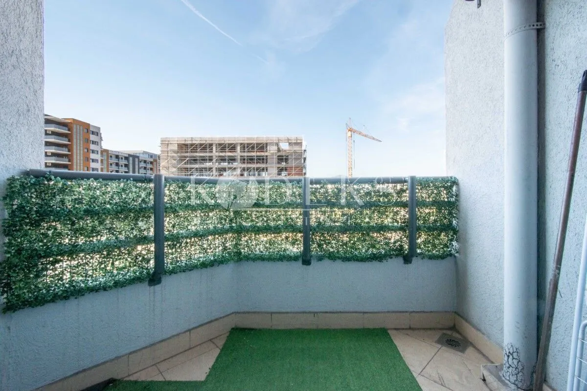 Manji balkon sa ogradom ukrašenom vještačkom travom i pogledom na okolne zgrade u izgradnji.