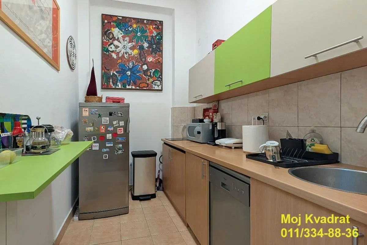 Manja kuhinja sa drvenim elementima, frižiderom i umetničkom slikom na zidu.