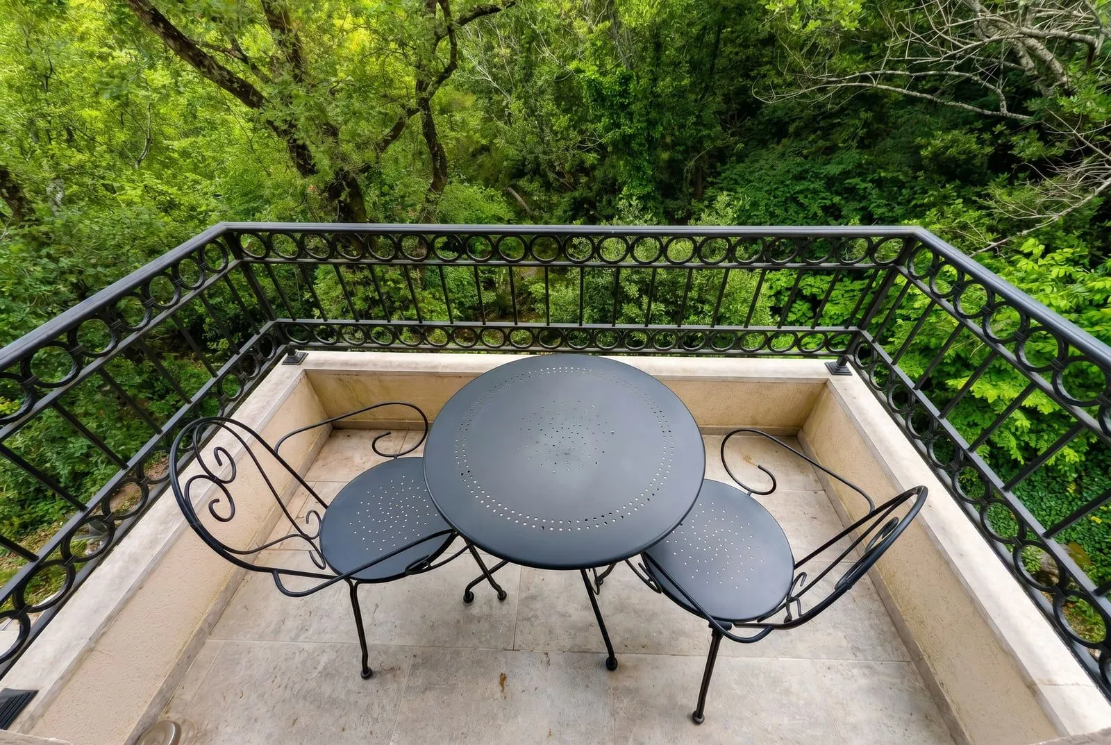 Balkon sa metalnim stolom i dvije stolice okružen zelenilom.
