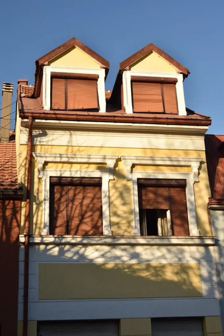 Dio fasade žute kuće sa prozorima i drvenim roletnama.