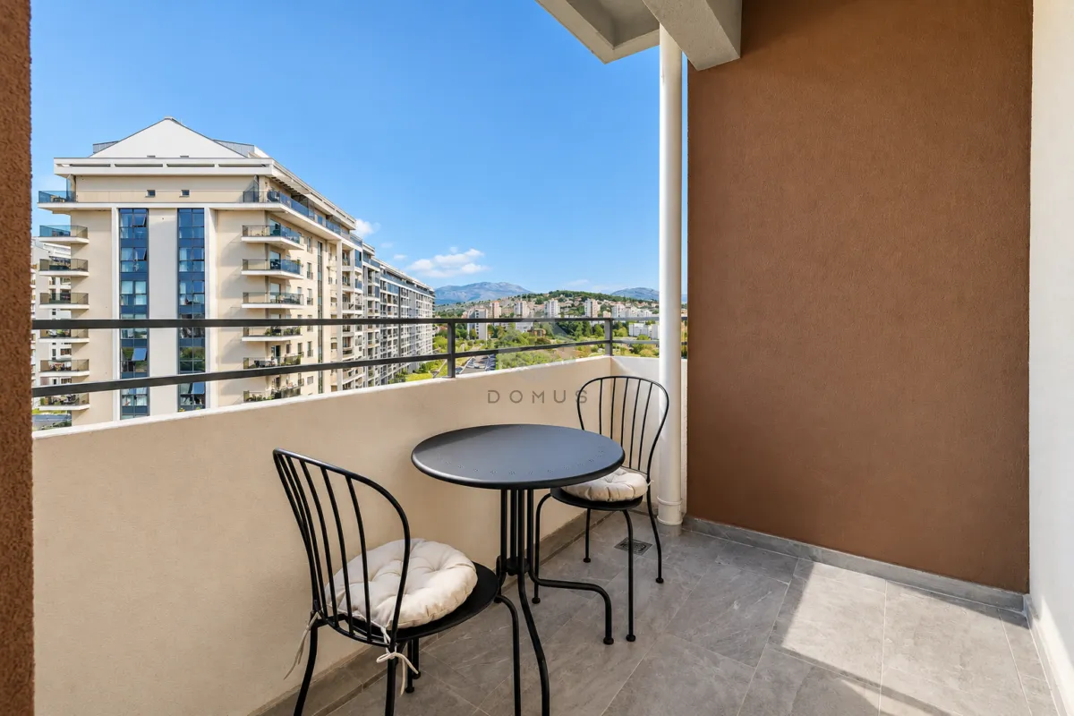 Natkriveni balkon sa garniturom za sjedenje i pogledom na okolne zgrade i brda.