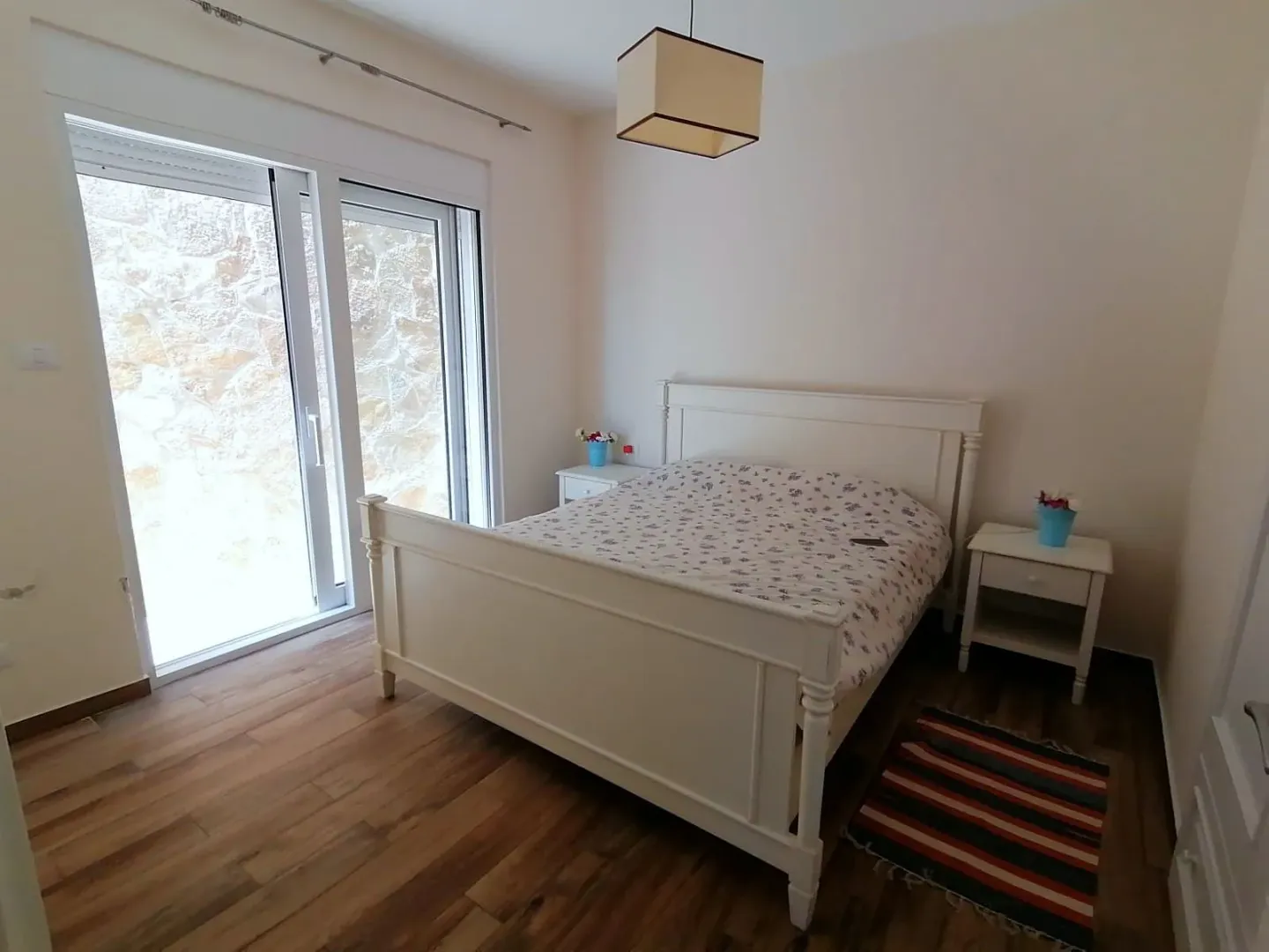 Spavaća soba sa bračnim krevetom, noćnim stočićima i balkonskim vratima.