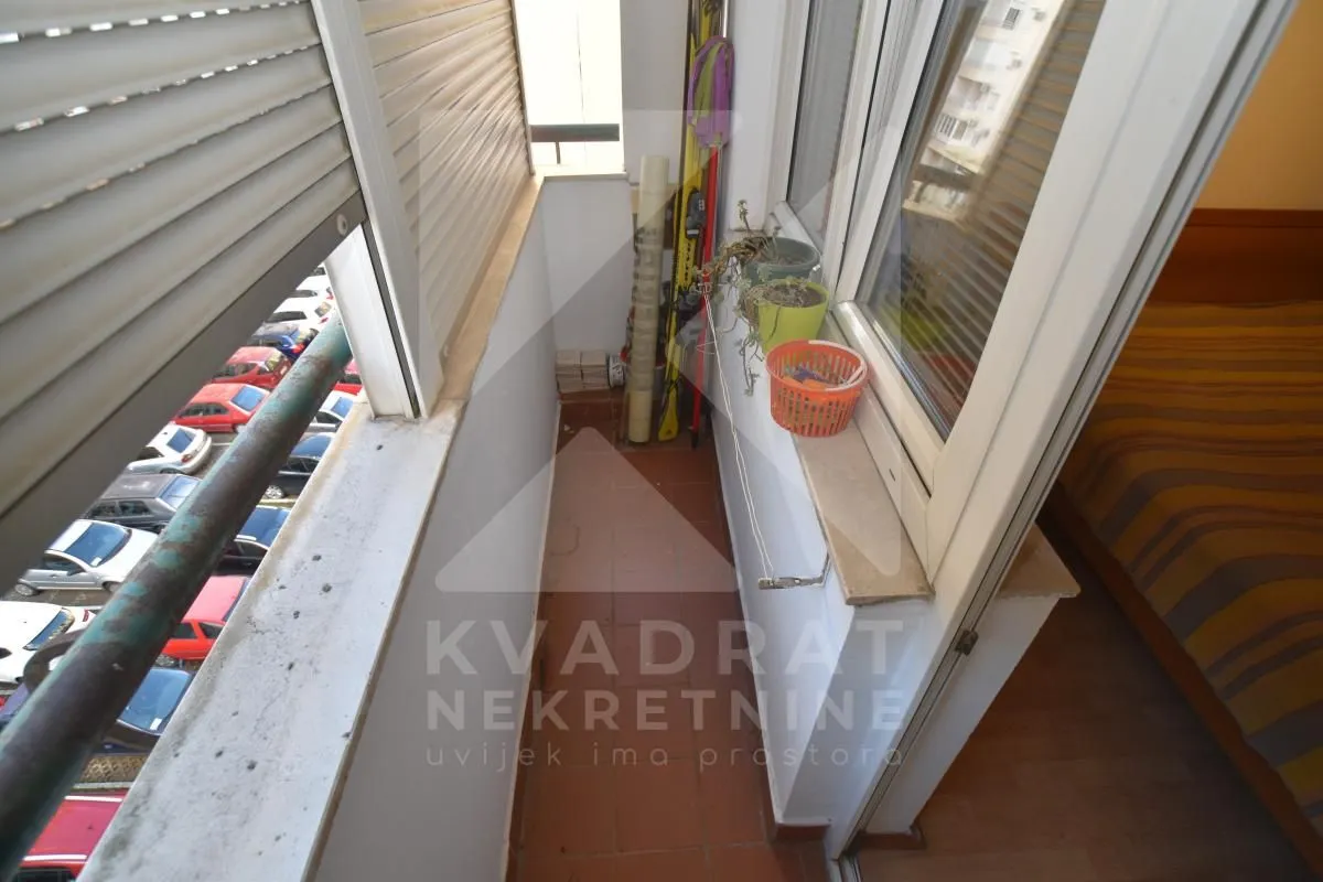 Uski zatvoreni balkon sa popločanim podom i pogledom na parking.