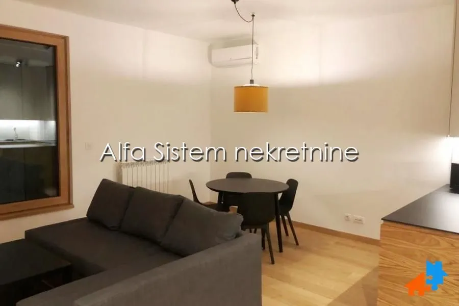 Dnevna soba sa ugaonom garniturom, trpezarijskim stolom i kuhinjskim elementima.