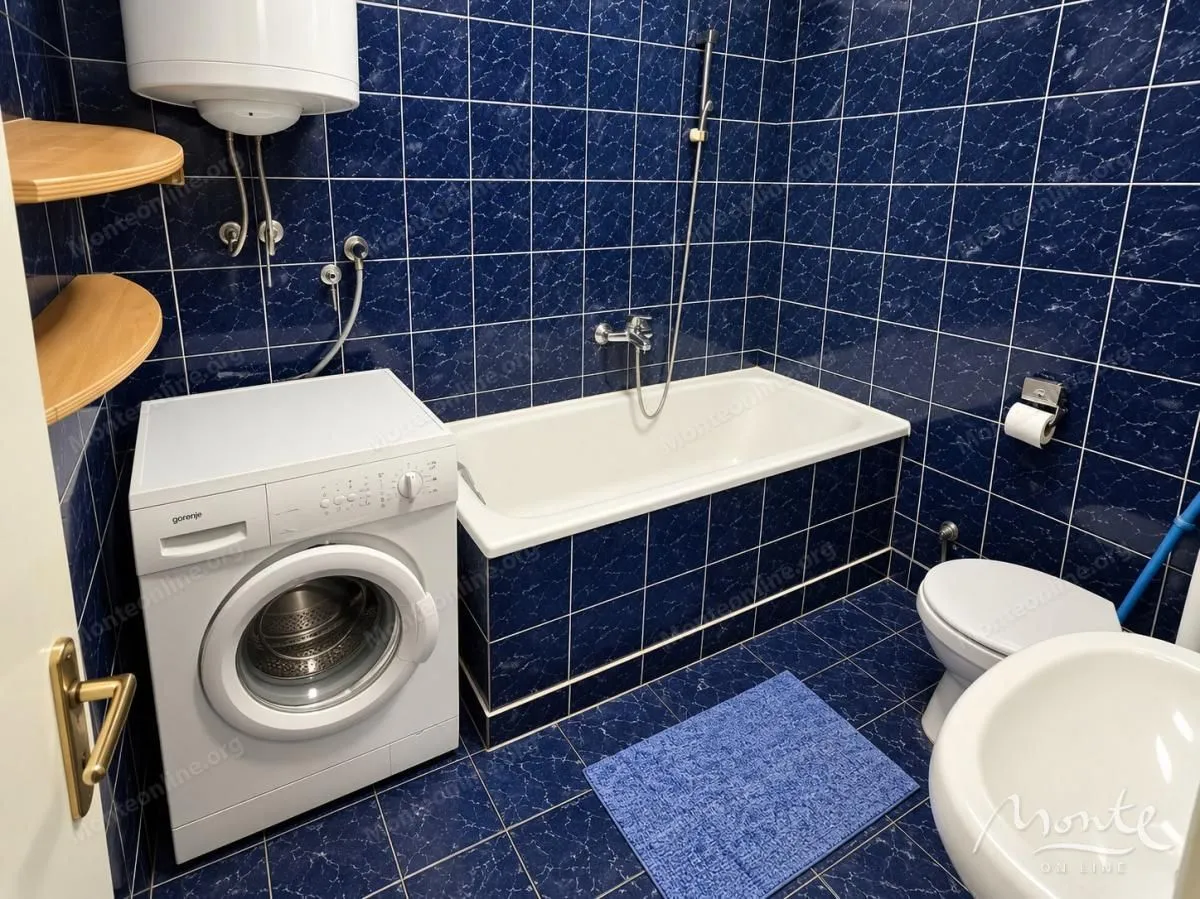 Kupatilo obloženo plavim pločicama sa kadom, veš mašinom i sanitarijama.