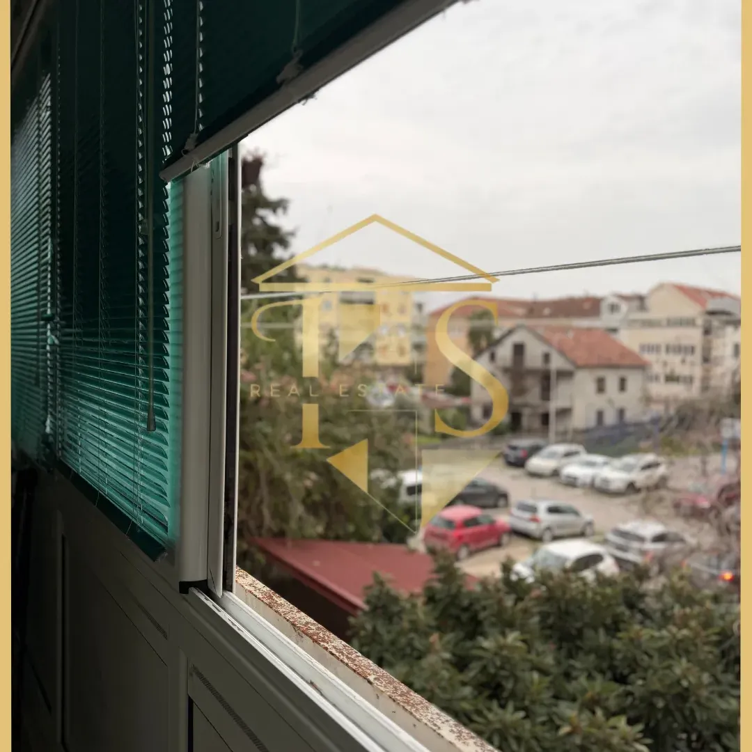 Pogled kroz prozor sa roletnama na okolne zgrade i parking.