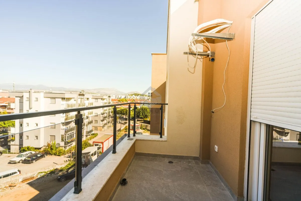 Nenamješten balkon sa metalnom ogradom i pogledom na okolne zgrade i naselje.