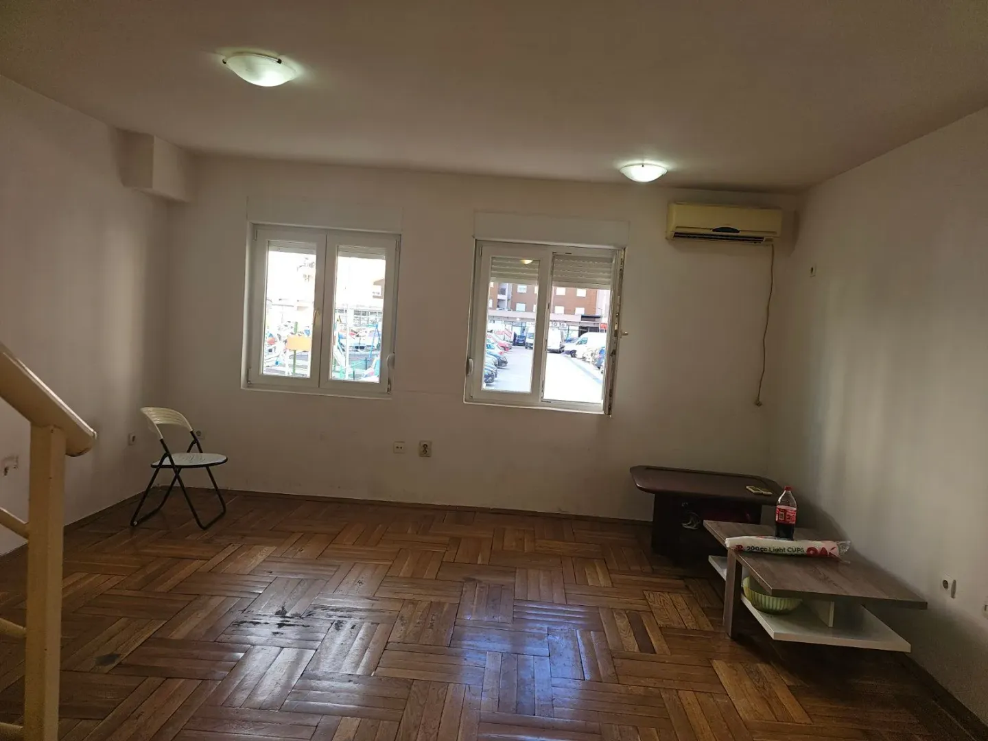 Empty living room with wooden parquet flooring, two windows, and an air conditioning unit.