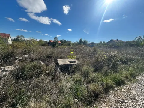 Pogled na zarasli plac sa betonskim šahtom i kućama u pozadini. - image 2