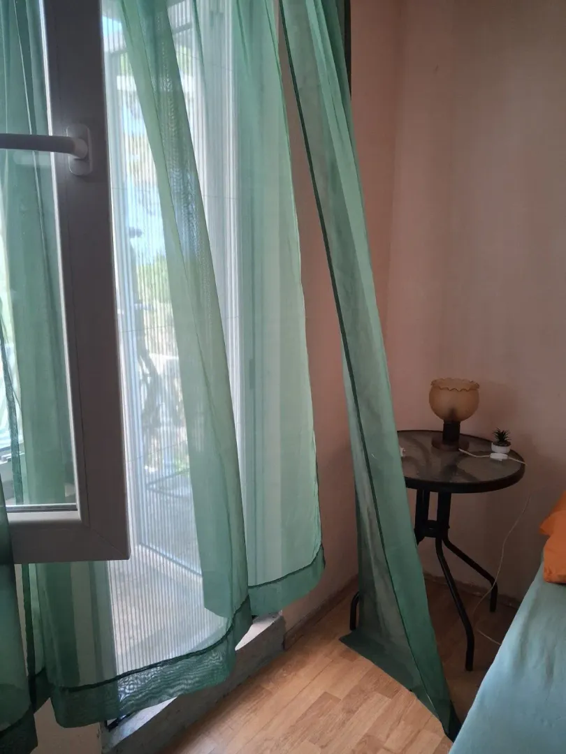 View of a window with green curtains and a small table with a lamp in the bedroom.