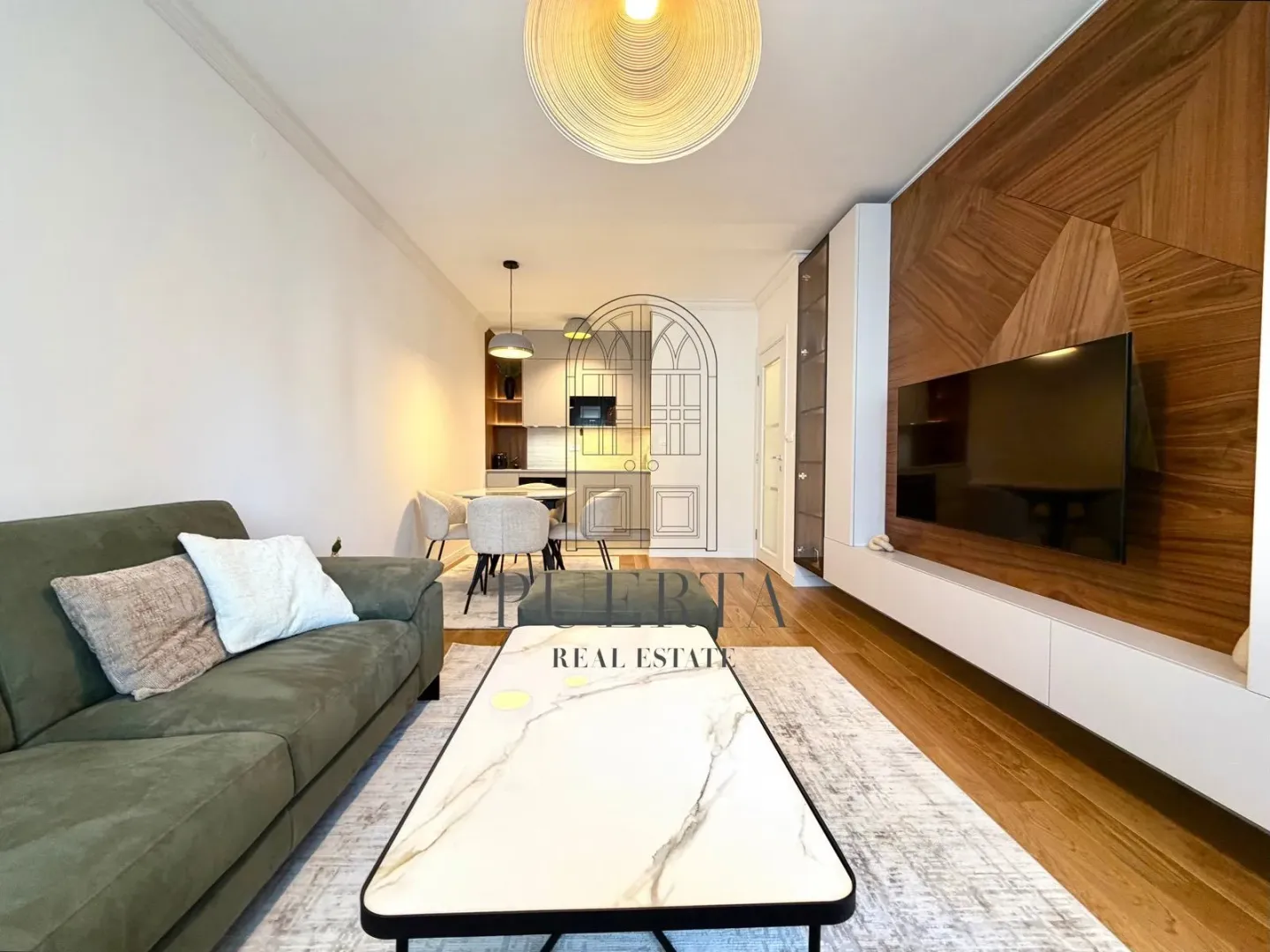 Modern living room with a sofa, TV panel, and dining area in the background.