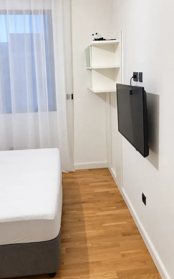 Svjetla spavaća soba sa krevetom, prozorom i zidnim televizorom.