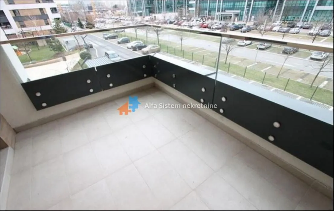 Prostrani popločani balkon sa staklenom ogradom i pogledom na parking i okolinu.