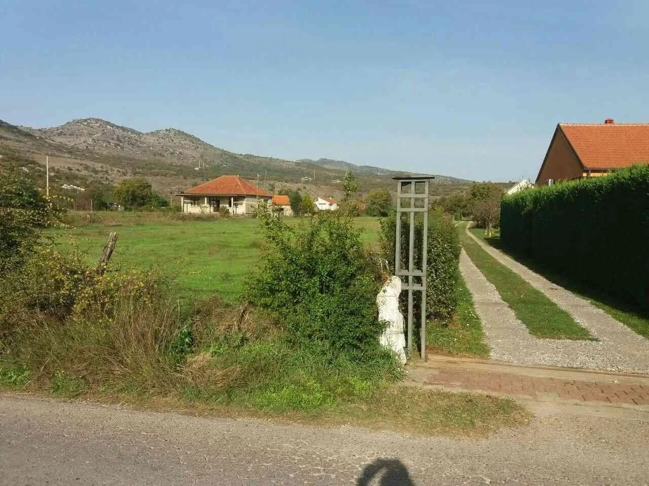 Pogled na prostrani plac sa prilaznim putem i kućama u pozadini.