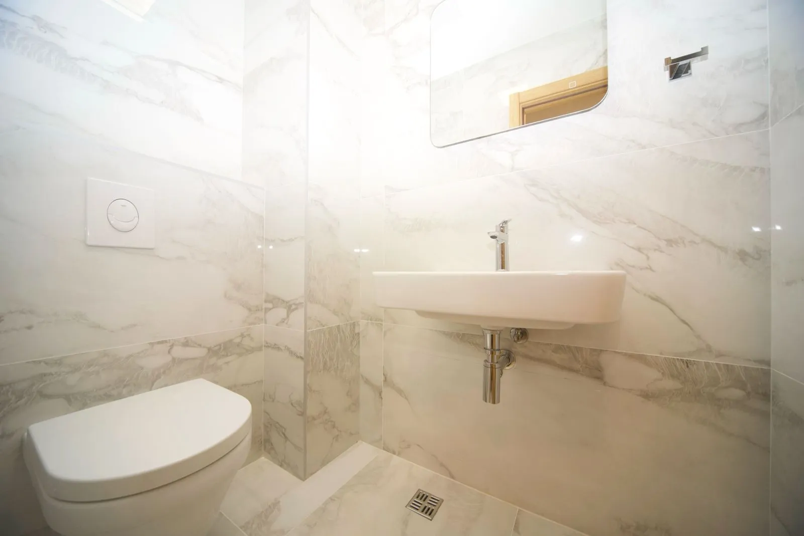 Smaller bathroom with a washbasin, toilet bowl, and mirror lined with marble tiles.