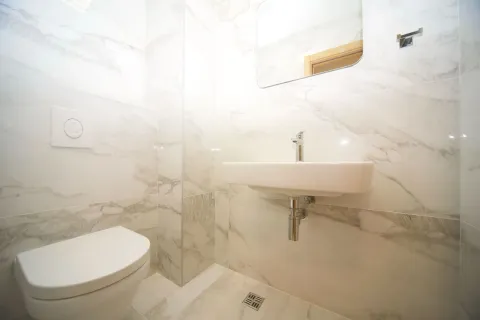 Smaller bathroom with a washbasin, toilet bowl, and mirror lined with marble tiles. - image 11