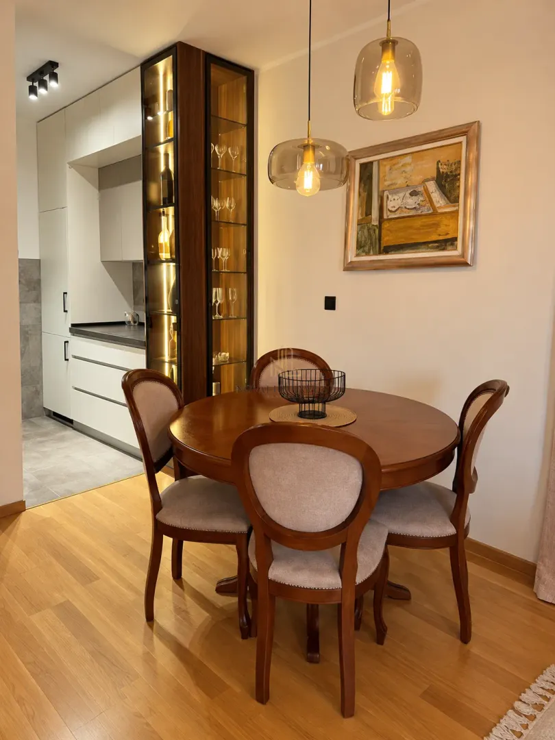 Dining area with a round wooden table and chairs, featuring an illuminated display cabinet and a view of the kitchen.