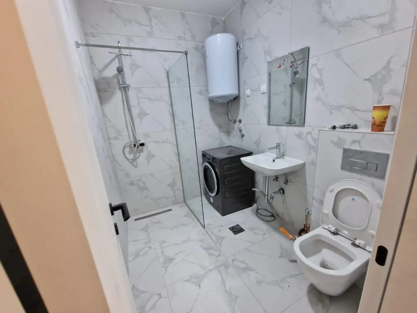 Moderne kupatilo obloženo keramičkim pločicama sa tuš kabinom, umivaonikom, WC šoljom i veš mašinom.