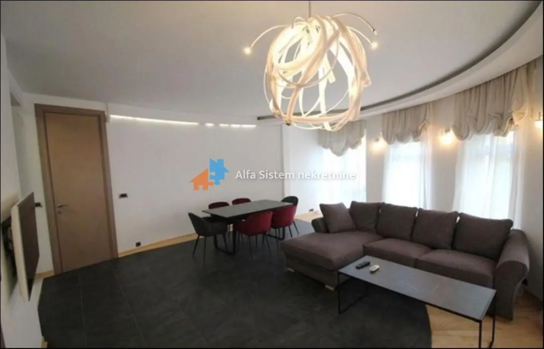 Prostrani dnevni boravak sa garniturom, trpezarijskim stolom i modernim lusterom.