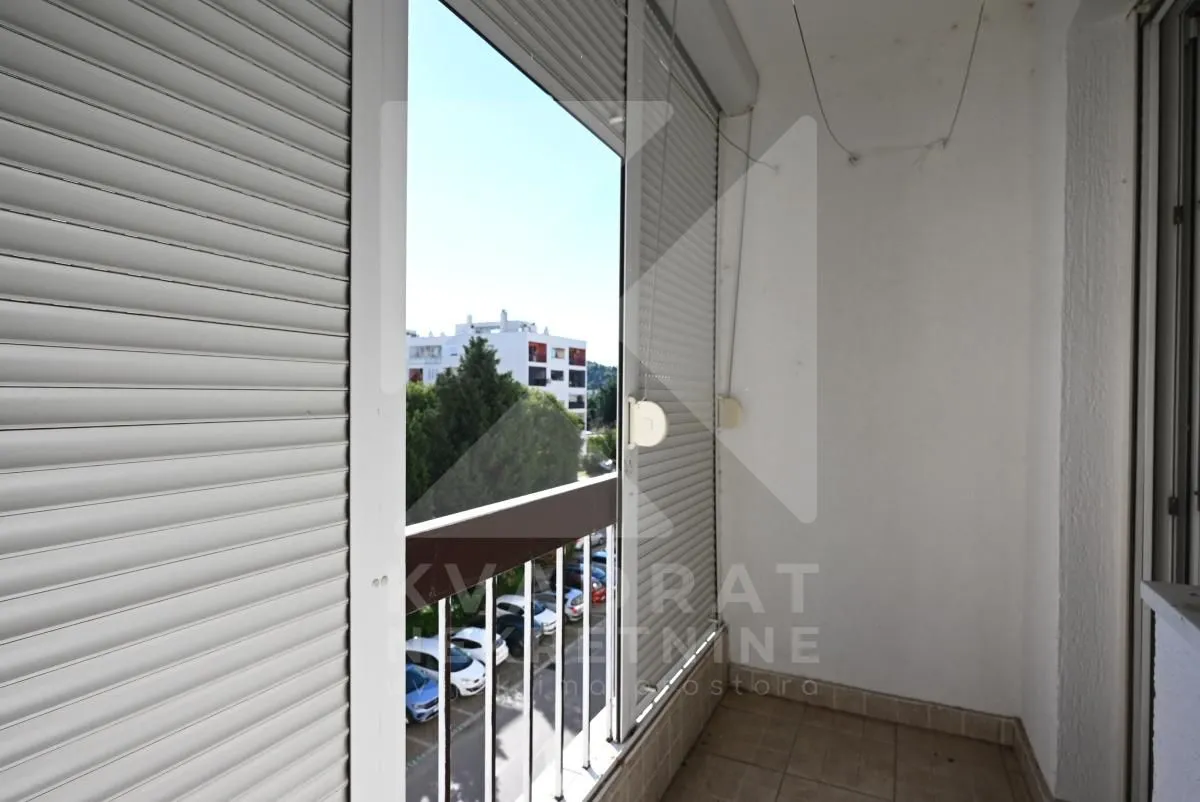 Covered balcony with roller shutters and a view of surrounding buildings and parking.