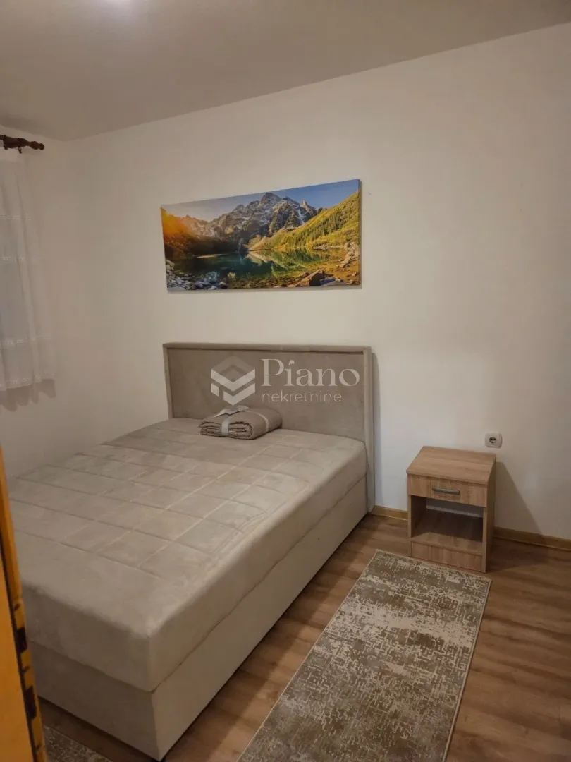 Spavaća soba sa bračnim krevetom, noćnim stočićem i slikom na zidu.
