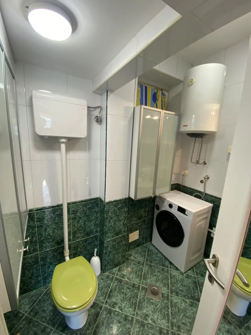 Manje kupatilo sa zelenim pločicama, wc šoljom i veš mašinom.
