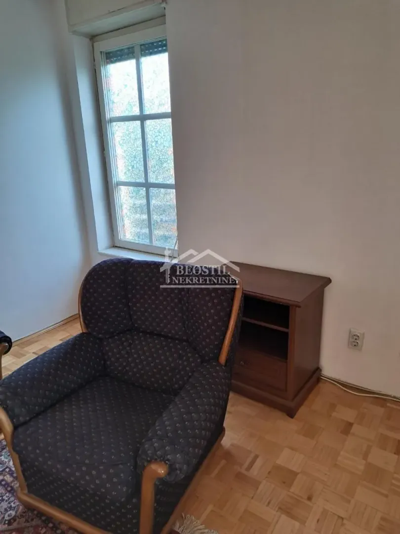 A small armchair and a wooden side table by the window in a room with parquet flooring.