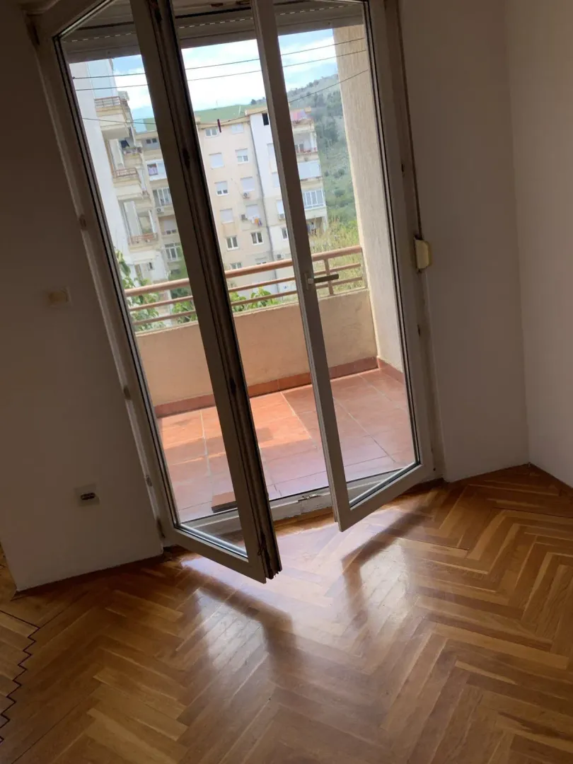 Prazna soba sa očuvanim parketom i otvorenim vratima koja vode na balkon.