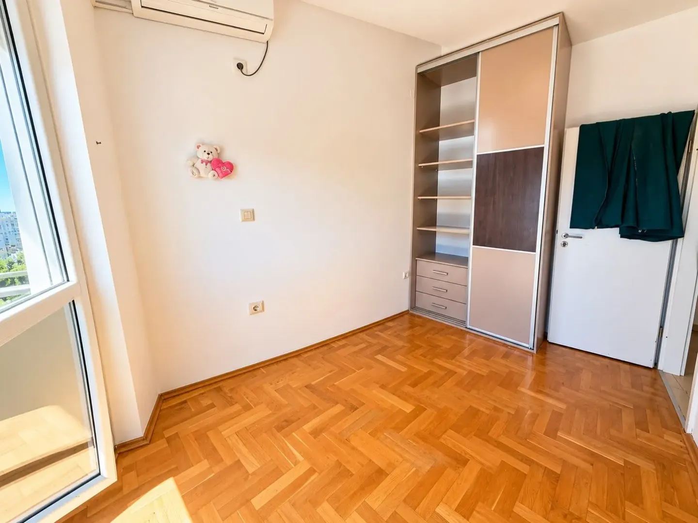 Prazna soba sa parketom, ugradnim ormarom i balkonskim vratima.