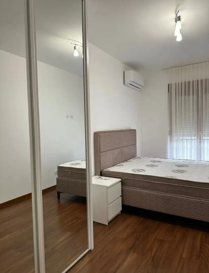 Svijetla spavaća soba sa bračnim krevetom, noćnim stočićem i velikim ogledalom.