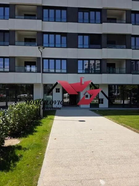 Pogled na fasadu i ulaz u stambenu zgradu sa popločanim stazama i uređenim travnjakom.