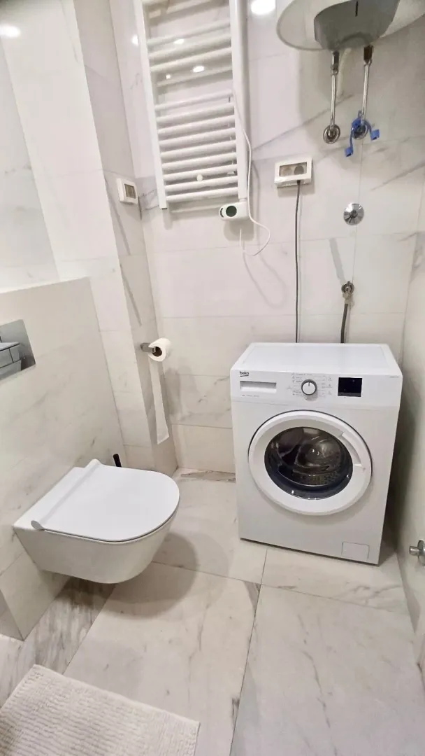 Moderne kupatilo sa bijelim pločicama, konzolnom šoljom i veš mašinom.