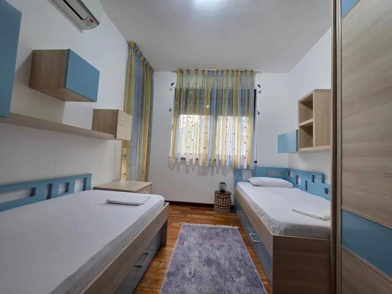 Children's bedroom with two beds, wooden flooring, and a window with yellow curtains.