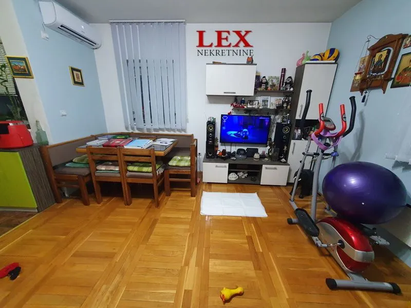 Dnevna soba sa garniturom za sedenje, trpezarijskim stolom i spravama za vežbanje.