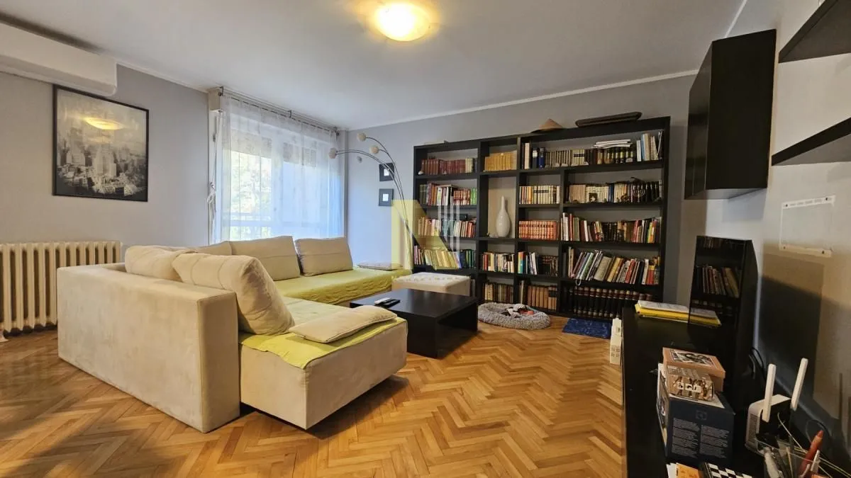 Dnevna soba sa ugaonom garniturom, velikom bibliotekom i parketom na podu.