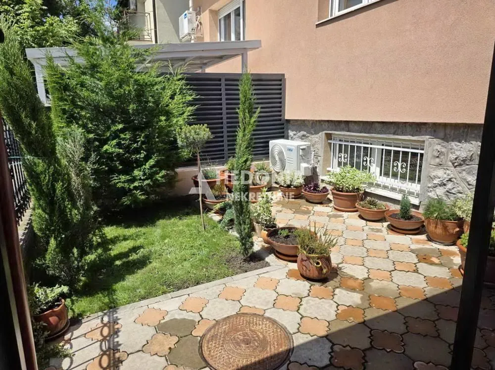 Popločano dvorište sa travnjakom i saksijama sa cvećem pored kuće.