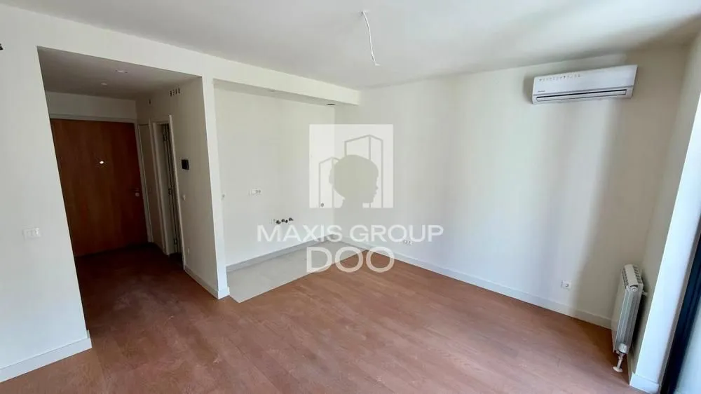 Empty living room with wooden flooring, air conditioning unit and kitchen connections.