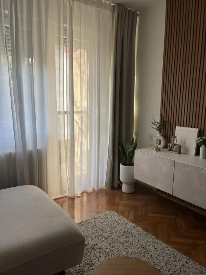Dio dnevne sobe sa bijelom komodom, drvenim panelima i velikim prozorom sa zavjesama.