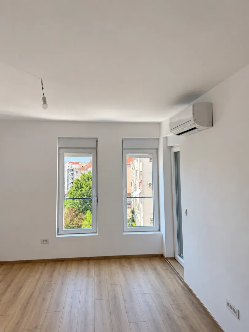 An empty room with parquet flooring, two windows, an air conditioner, and balcony doors.