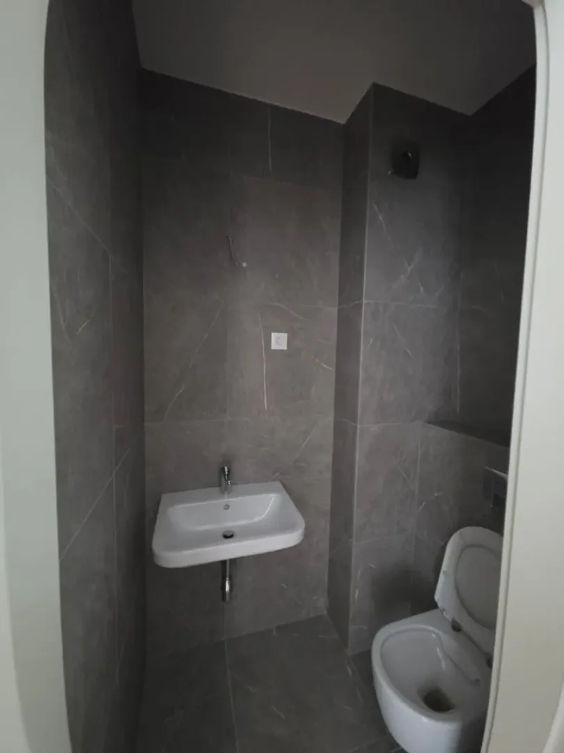 Manje kupatilo sa umivaonikom i šoljom, obloženo sivim pločicama.