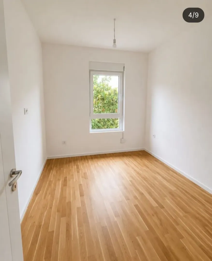 Prazna soba sa bijelim zidovima, drvenim parketom i prozorom.