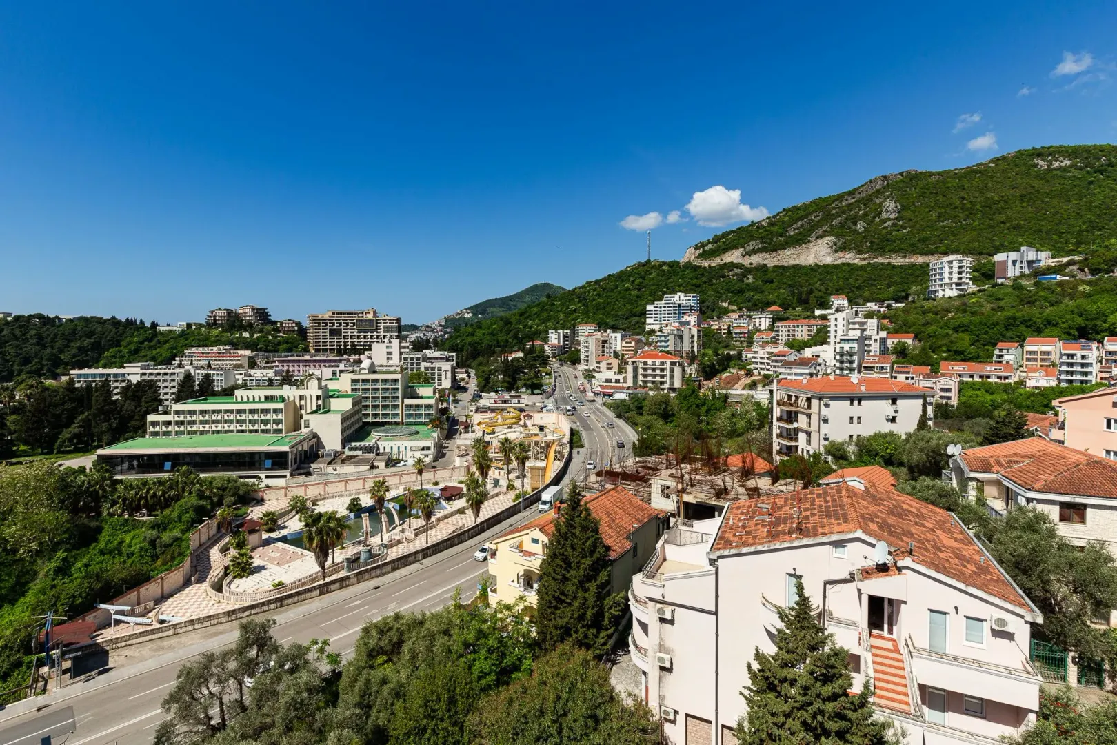 Panoramski pogled sa visine na naselje, hotele i okolna brda pod plavim nebom.