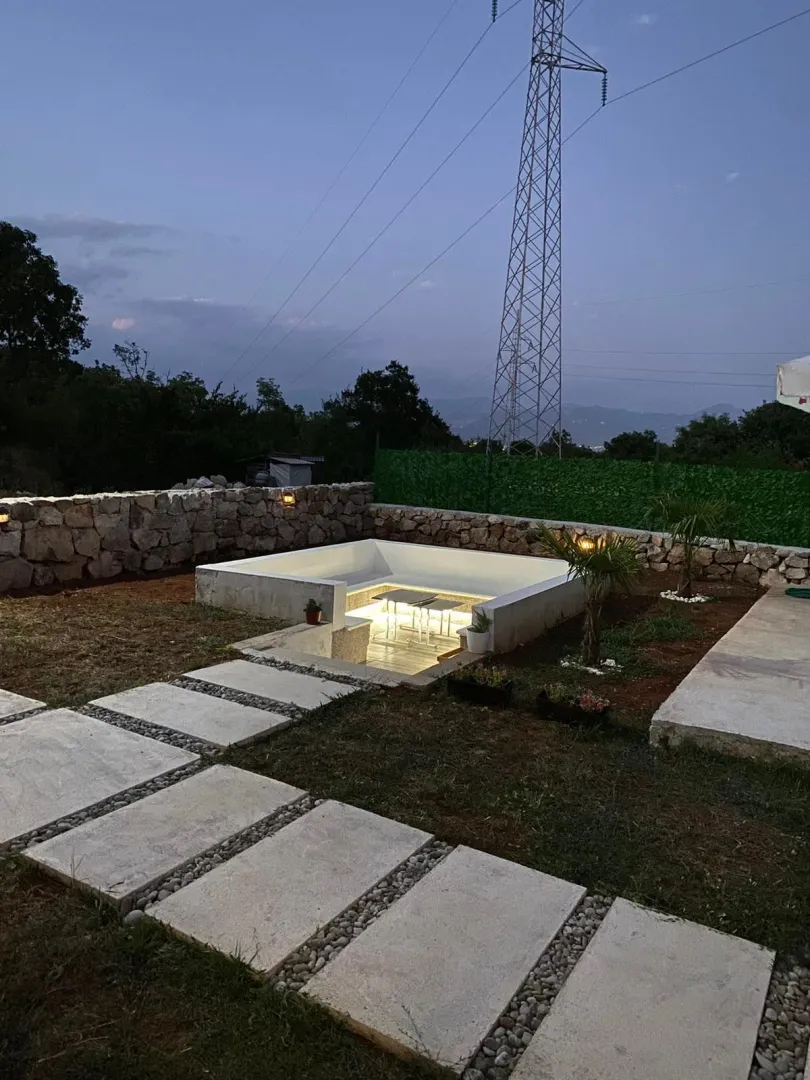 Yard with a paved walkway, illuminated sunken seating area, and stone wall at dusk.