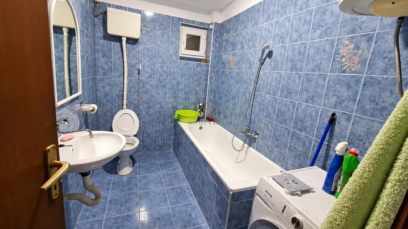Kupatilo obloženo plavim pločicama sa kadom, umivaonikom, toaletom i veš mašinom.