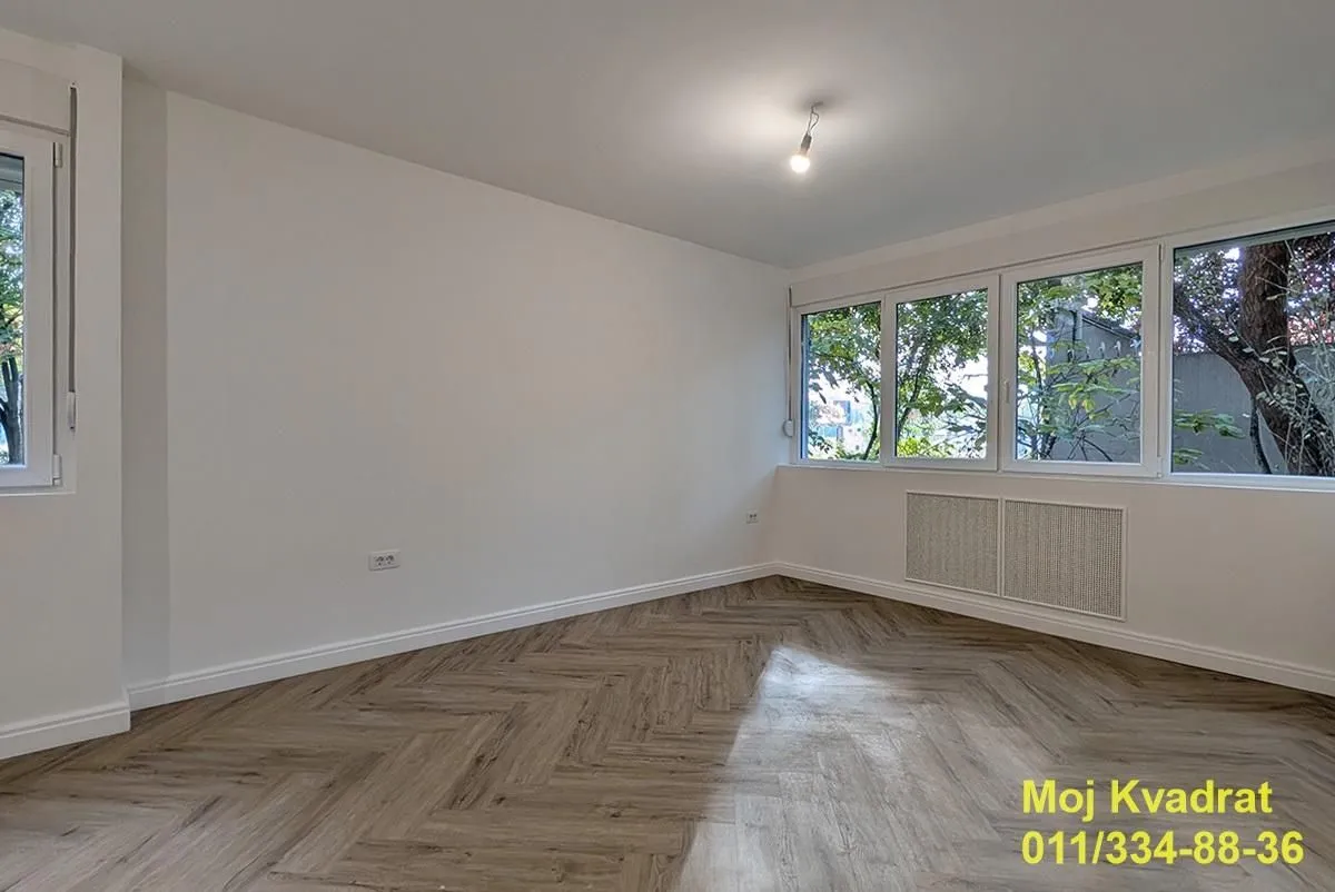 Prazna soba sa belim zidovima, drvenim podom i velikim prozorima.